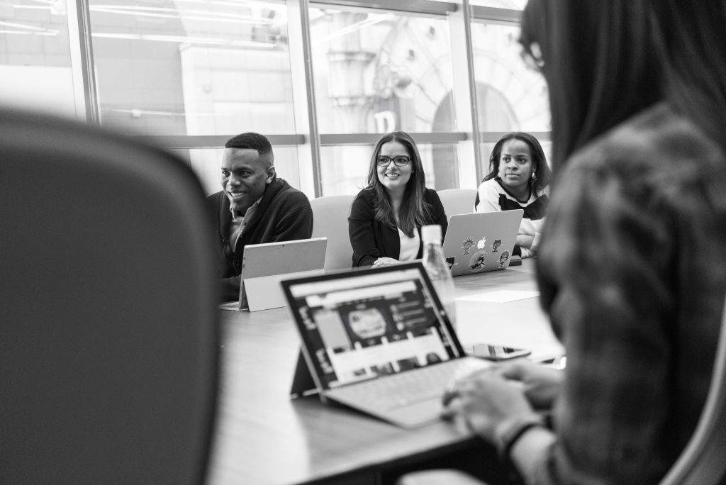 Drei Mitarbeitende sitzen lächelnd an einem Konferenztisch mit Laptops und Tablets während eines Meetings