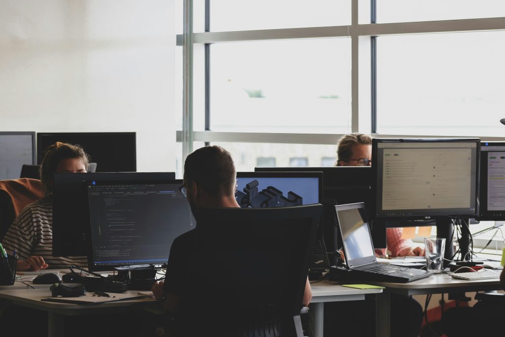 Drei Personen arbeiten in einem hellen Großraumbüro an mehreren Bildschirmen mit Code- und 3D-Anwendungen