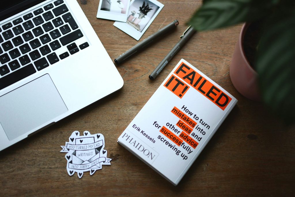 Buch 'Failed It!' von Erik Kessels auf einem Holztisch neben einem MacBook, Stiften und Polaroid-Fotos