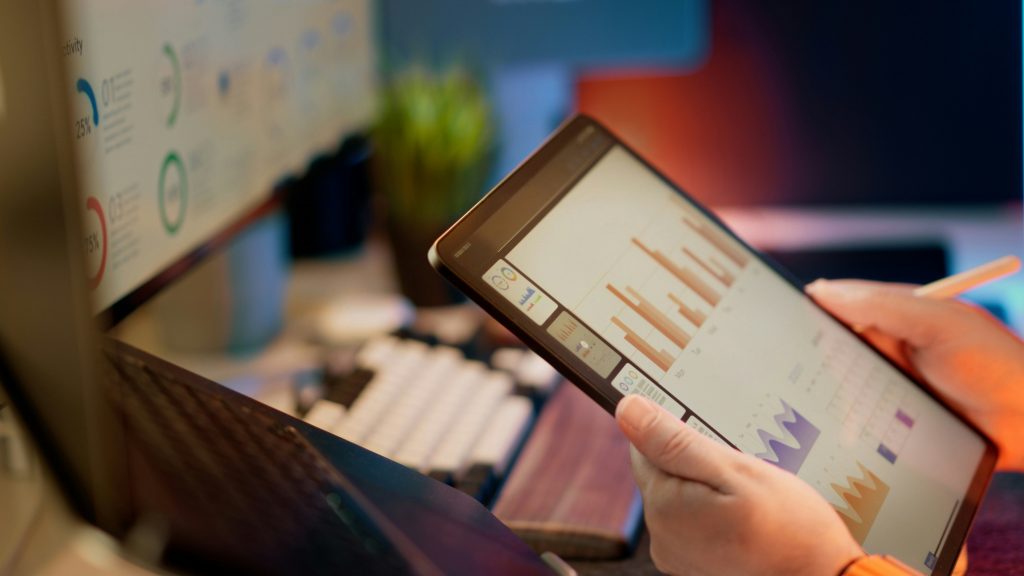 Hände halten ein Tablet mit Balken- und Flächendiagrammen, im Hintergrund ein Infografik-Poster im Büro