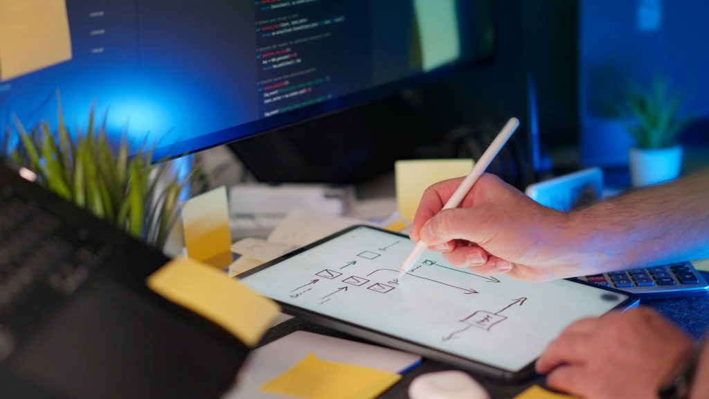 Hand mit Apple Pencil skizziert ein UI-Wireframe auf einem Tablet, im Hintergrund ein Monitor mit Code