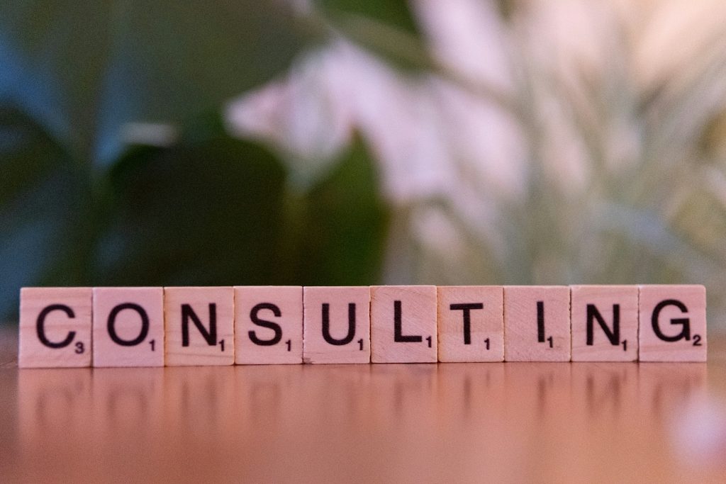 Scrabble-Buchstabensteine bilden das Wort CONSULTING auf einer Holzoberfläche vor unscharfem Pflanzenhintergrund