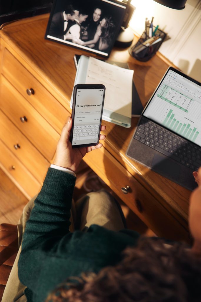 Person nutzt Microsoft Copilot auf dem Smartphone neben Laptop mit Excel Tabelle